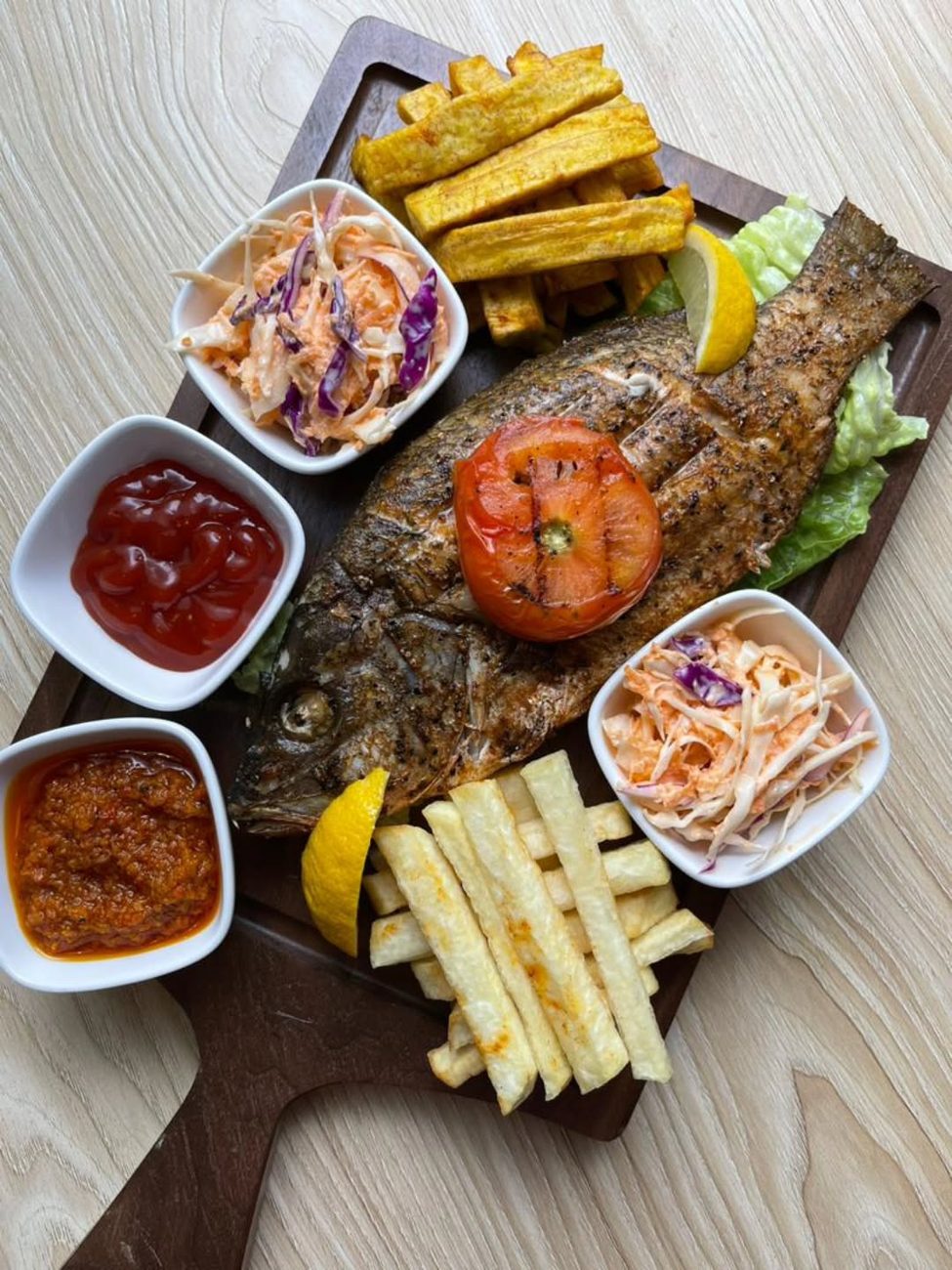 Grilled whole fish on a board with plantain, yam fries, coleslaw and dips